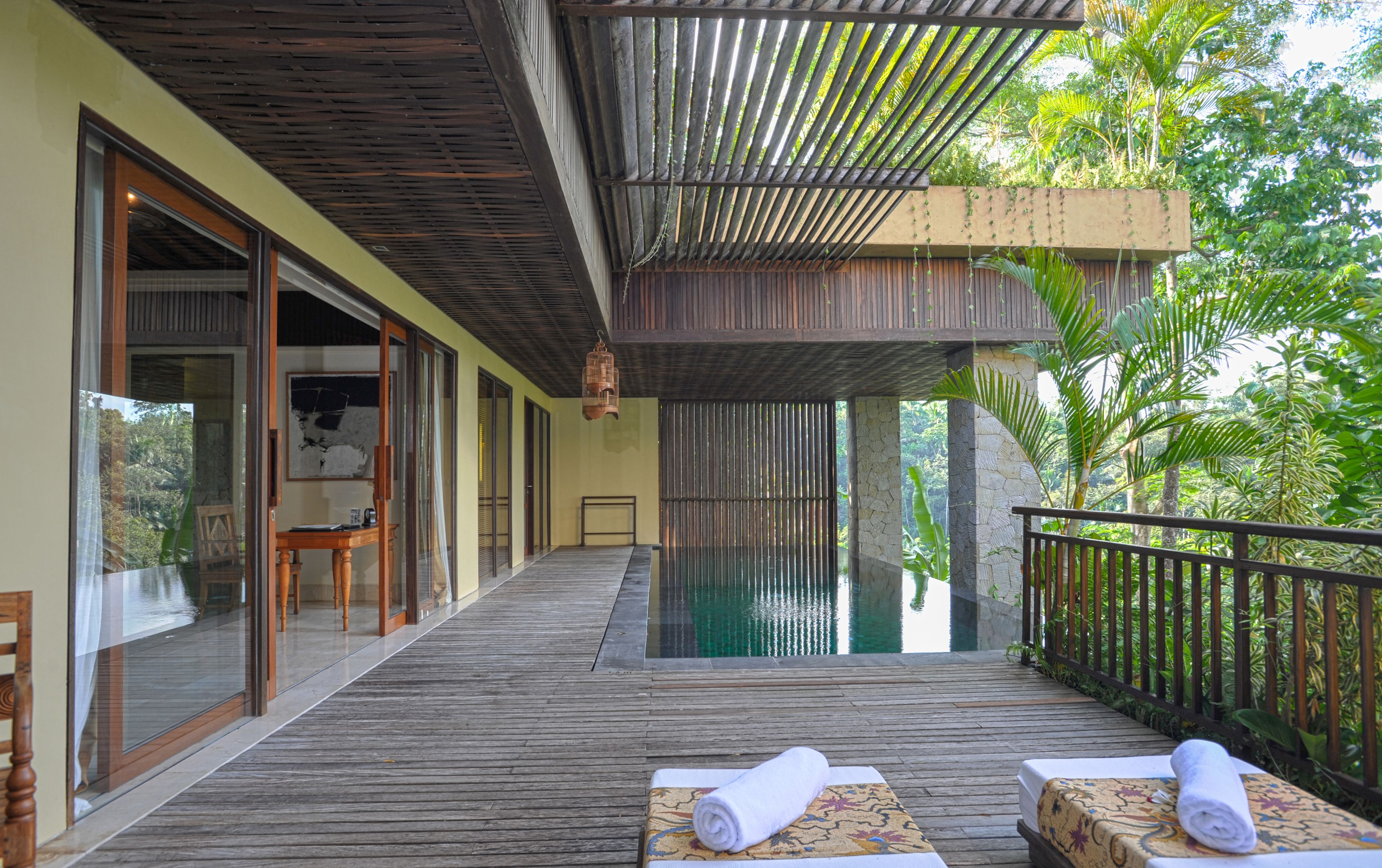 A wooden deck with a pool, wooden furniture, and a view of lush greenery.
