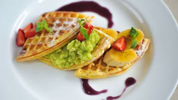 Un plato con waffles cubiertos de guacamole, fresas y rodajas de plátano, decorado con hojas de menta y bañado en salsa morada.