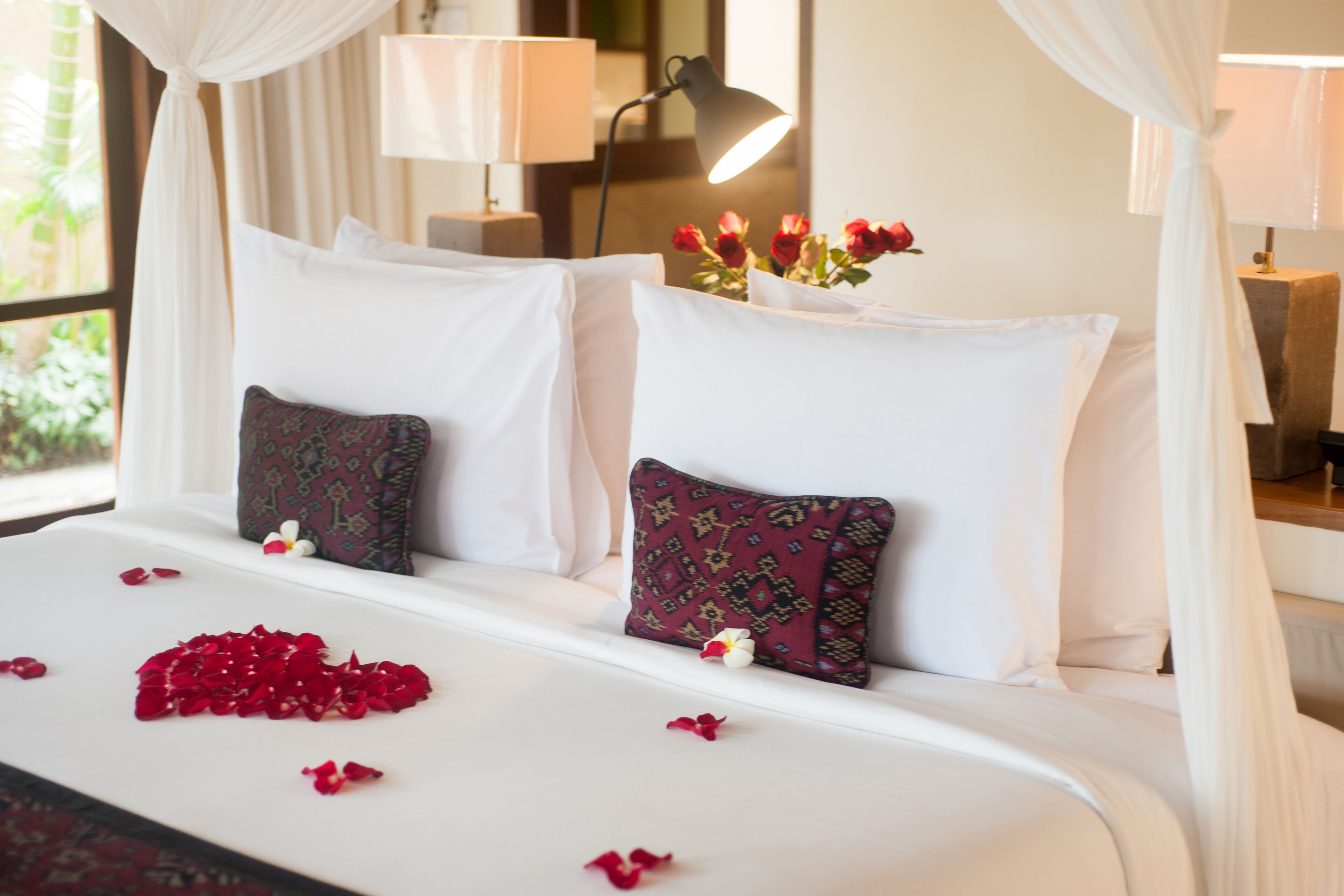 Una cama cuidadosamente tendida con sábanas blancas, pétalos de rosa roja y un jarrón de rosas rojas.