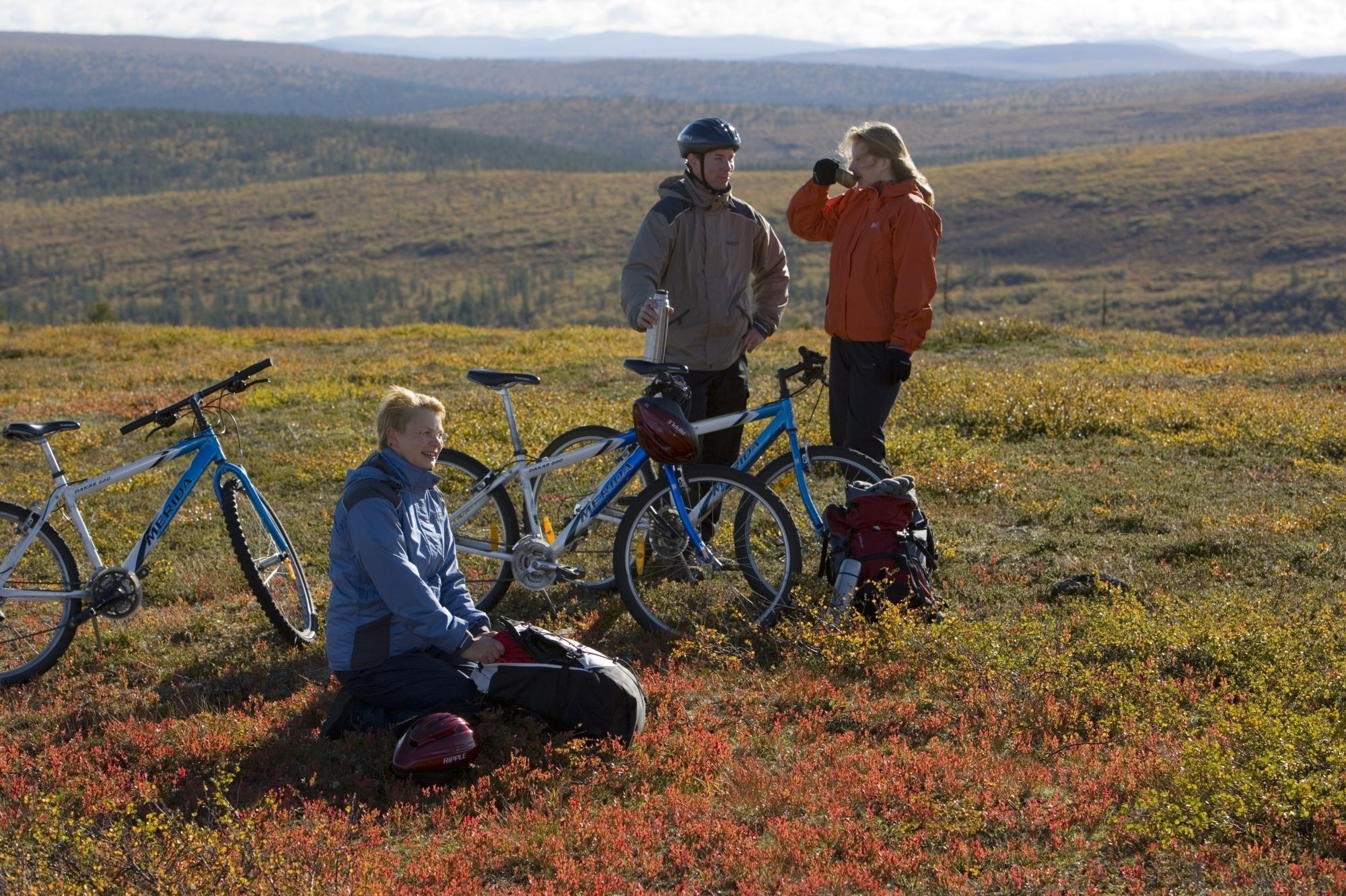 Trois personnes sont assises sur un champ d’herbe, à côté de leurs vélos, et sont entourées d’un paysage montagneux.