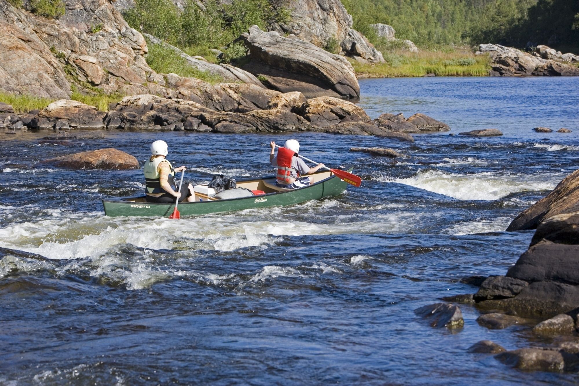 Dos personas en una canoa verde sobre un río, con rocas y vegetación al fondo.