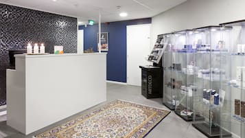 Zona de recepción con pared de mosaicos, mostrador de recepción, alfombra estampada y vitrina de vidrio con productos.