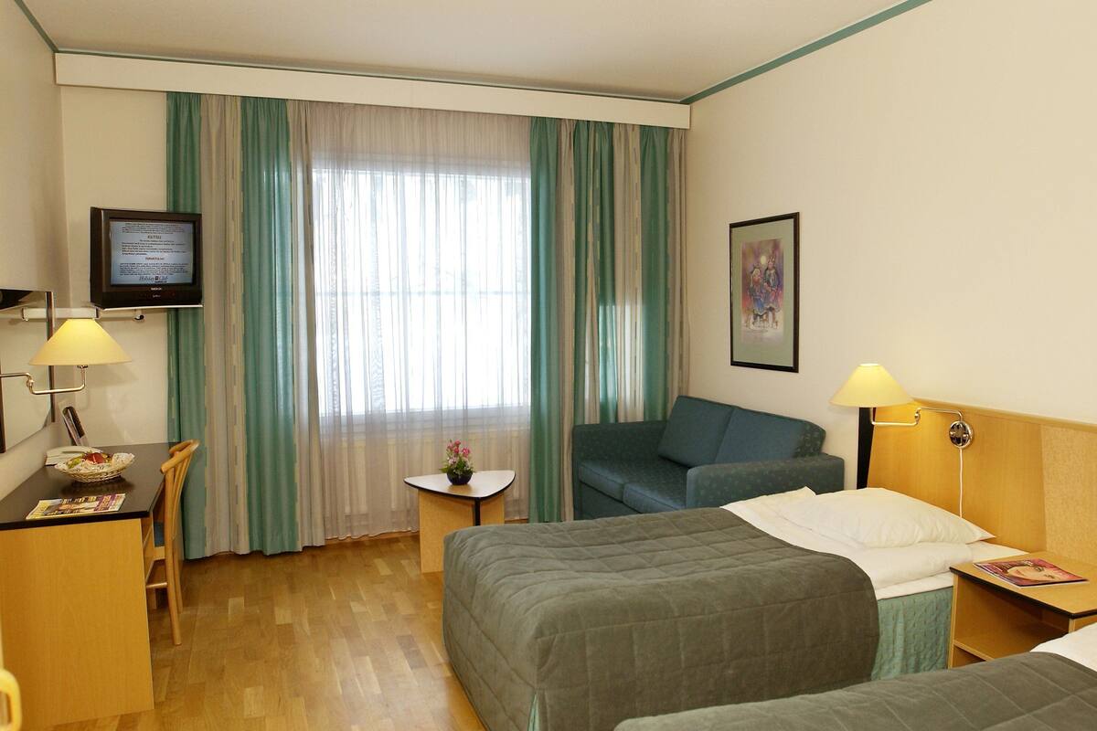 A hotel room with a bed, a desk with a lamp, a chair, a small table with a flower, and a television on the wall.