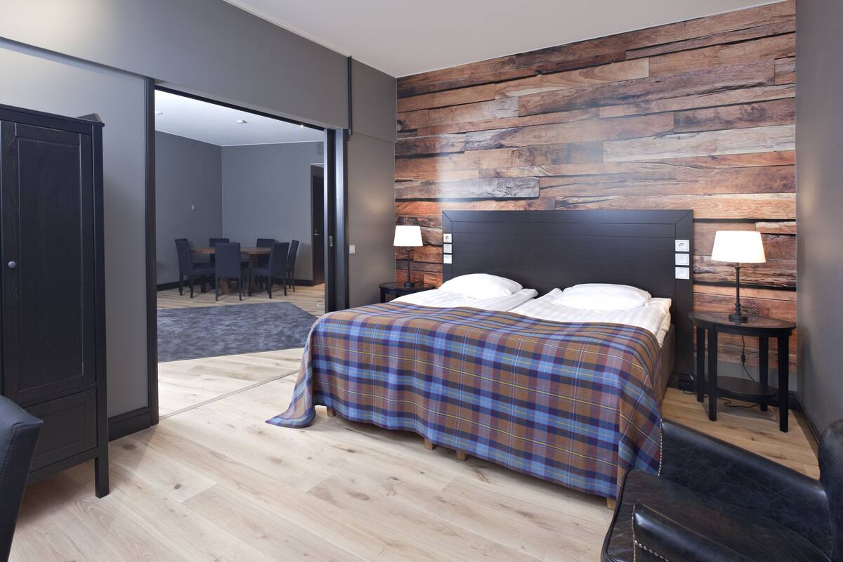 A modern hotel room with a large bed, two bedside tables with lamps, a wooden accent wall, a dark wardrobe, and a dining area visible through an open door.