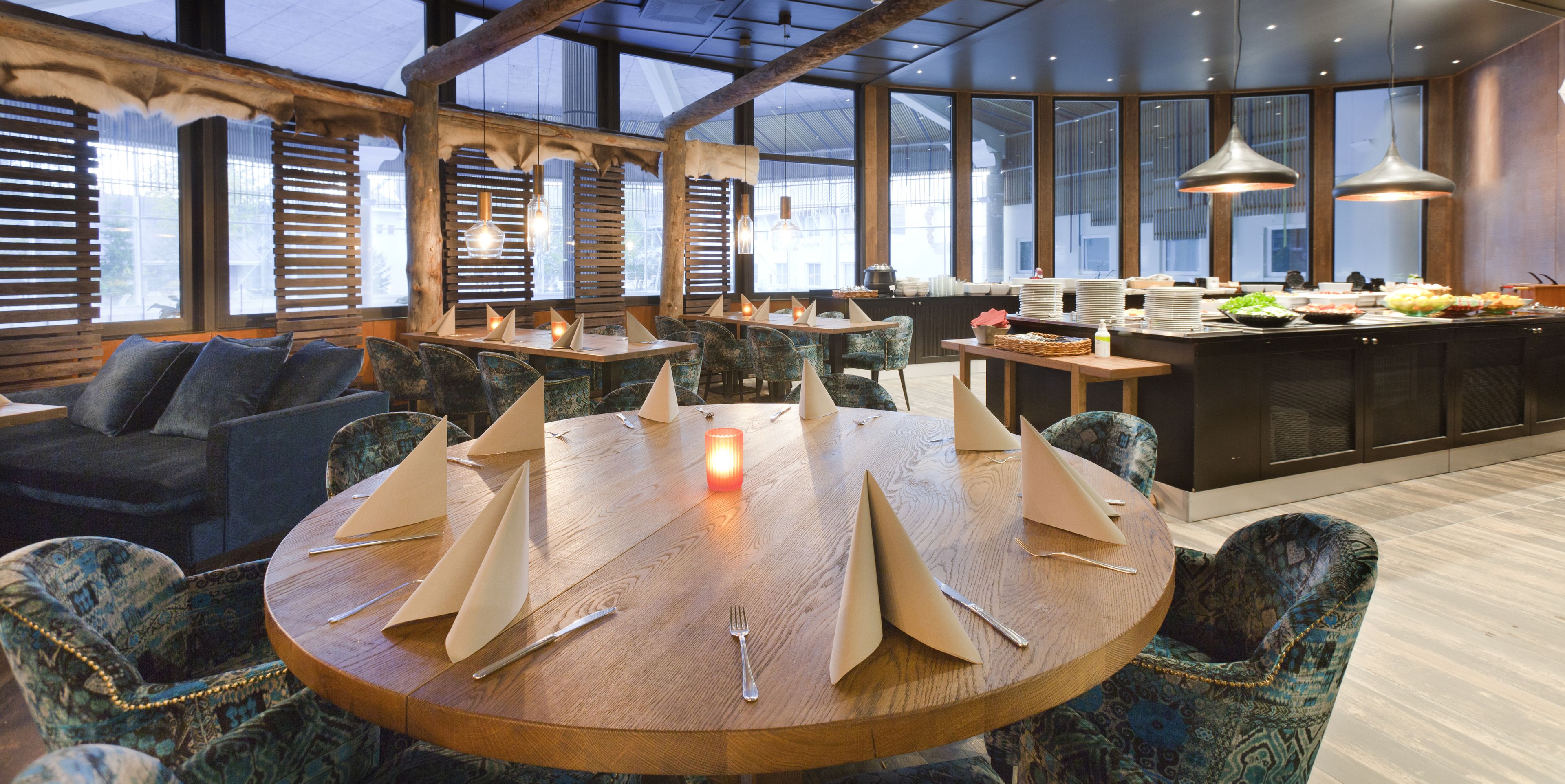 Um restaurante com mesas redondas preparadas para refeições, vigas de madeira no teto e grandes janelas.