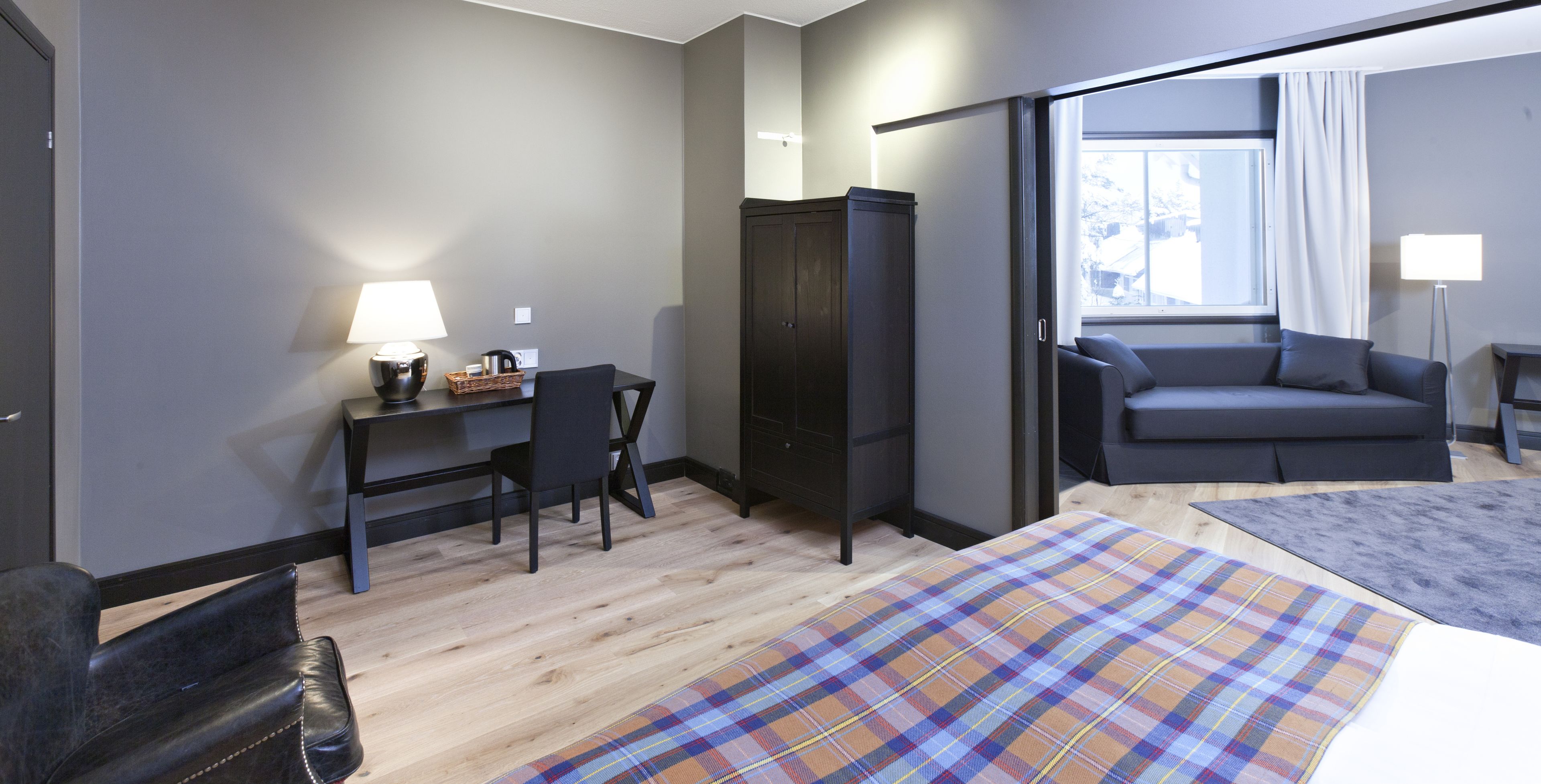A modern hotel room with a wooden floor, a bed with a plaid bedspread, a dark sofa, a desk with a lamp, and a wardrobe.
