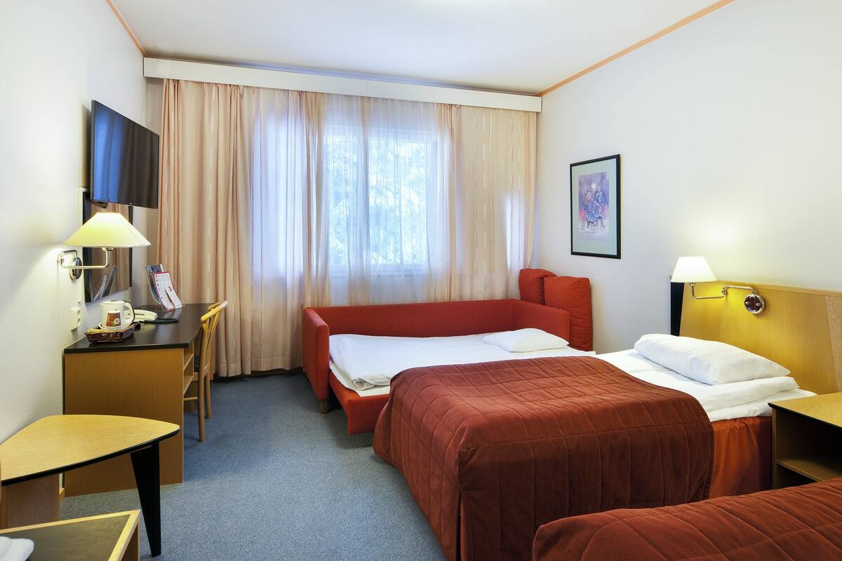 A hotel room with two beds, a desk, a chair, a TV, and a window with curtains.