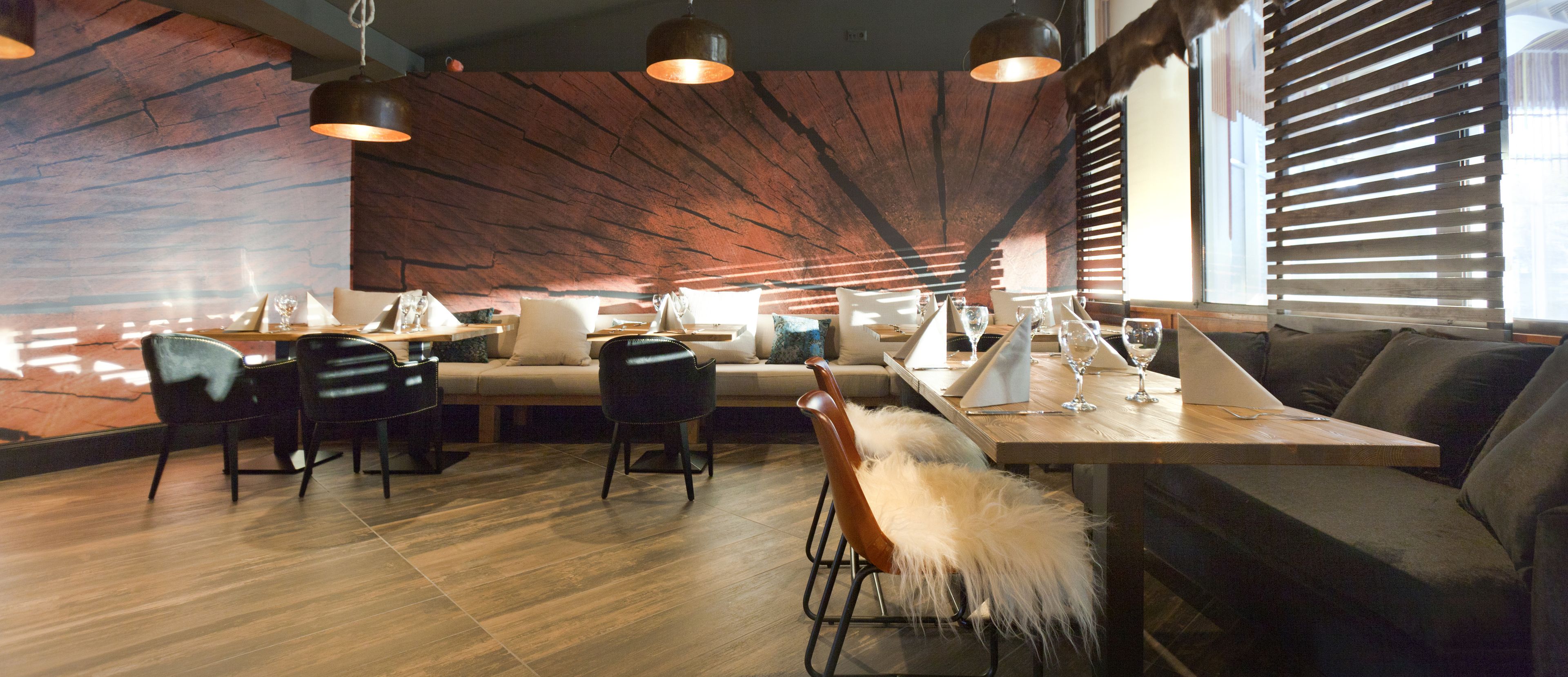 A modern dining area with wooden flooring, a wooden table, and black chairs. There is a sofa and a window with blinds in the background.
