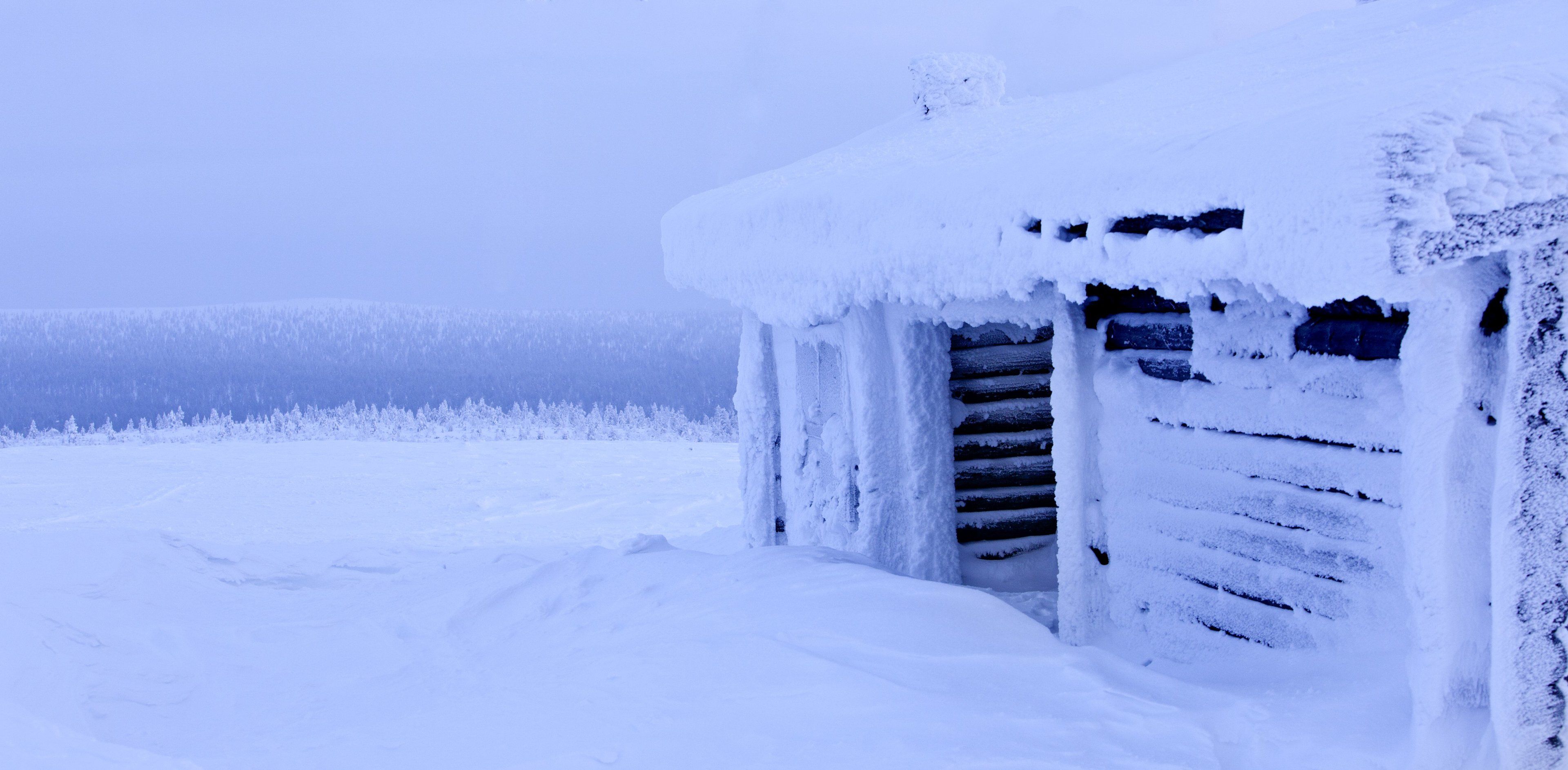 A snow-covered wooden cabin with a door and windows, surrounded by a snowy landscape.