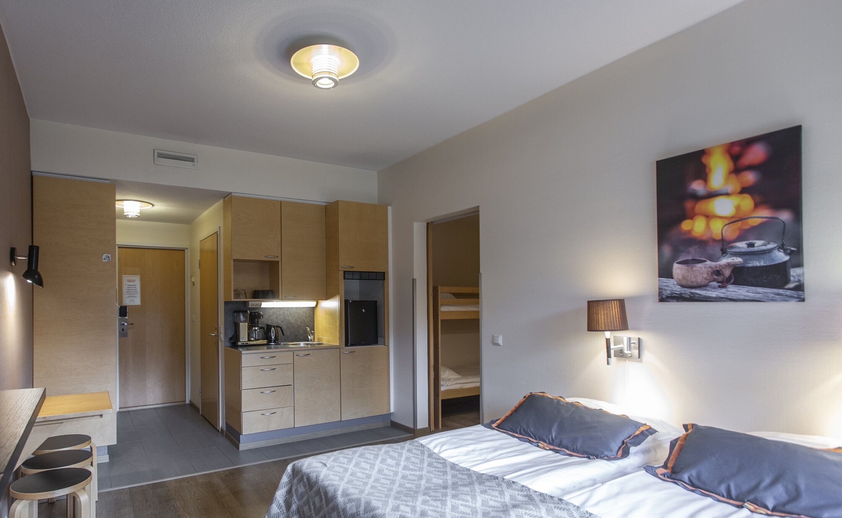 Une chambre d’hôtel avec un lit, des lampes de chevet, un tableau, une kitchenette et une salle de bain visible à travers une porte ouverte.