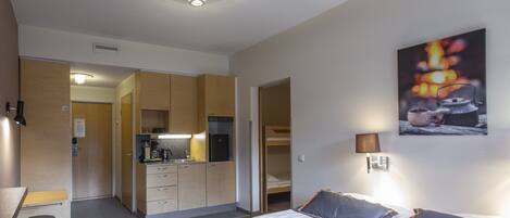 Une chambre d’hôtel avec un lit, des lampes de chevet, un tableau, une kitchenette et une salle de bain visible à travers une porte ouverte.