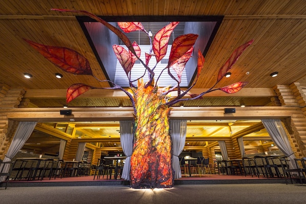 A large indoor space with a central decorative tree structure, wooden log walls, and tables and chairs arranged for dining.