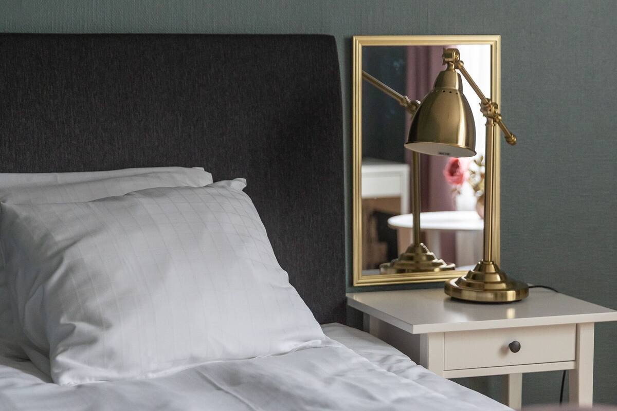 Un lit bien fait, avec du linge de lit blanc et des coussins roses, une table de chevet surmontée d’une lampe dorée, et un miroir qui reflète une partie de la pièce.