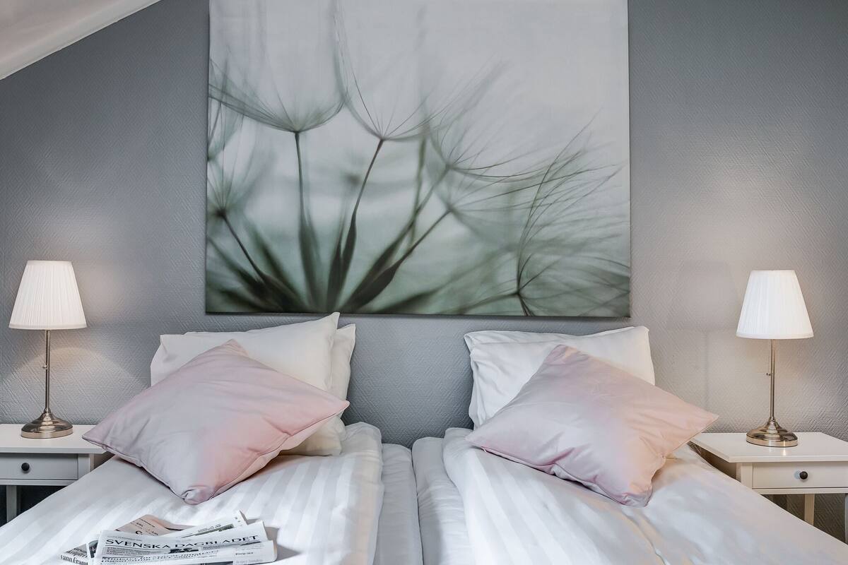 Un lit bien fait, avec une literie blanche et des coussins roses, flanqué de deux tables de chevet sur lesquelles reposent des lampes, le tout adossé à un mur gris à texture.