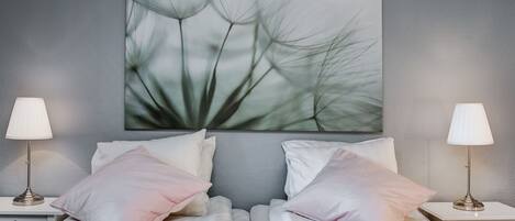 Una cama bien hecha con ropa blanca y almohadas rosadas, flanqueada por dos mesitas de noche con lámparas, frente a una pared gris con textura.