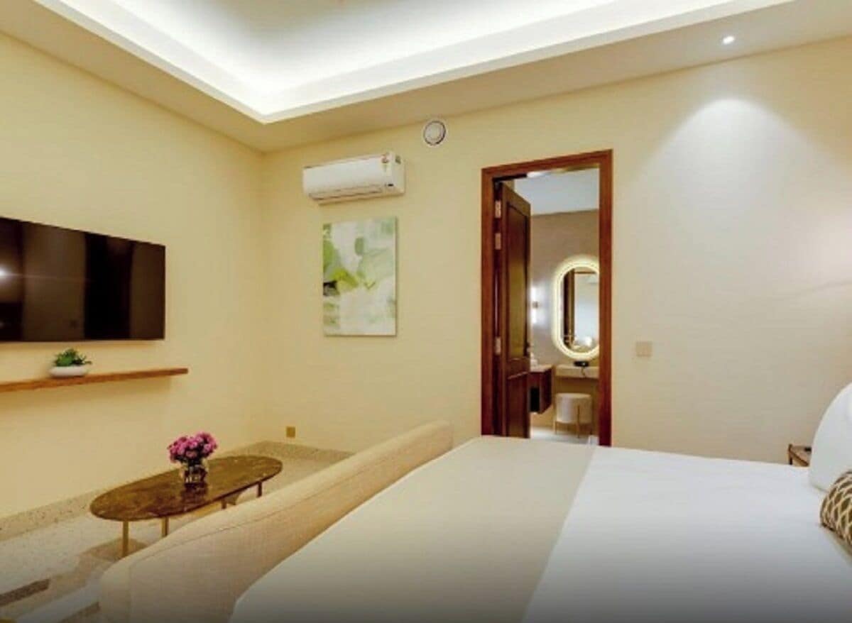 A hotel room with a bed, a small round table with a vase of flowers, a mounted TV, and a wall-mounted air conditioner.