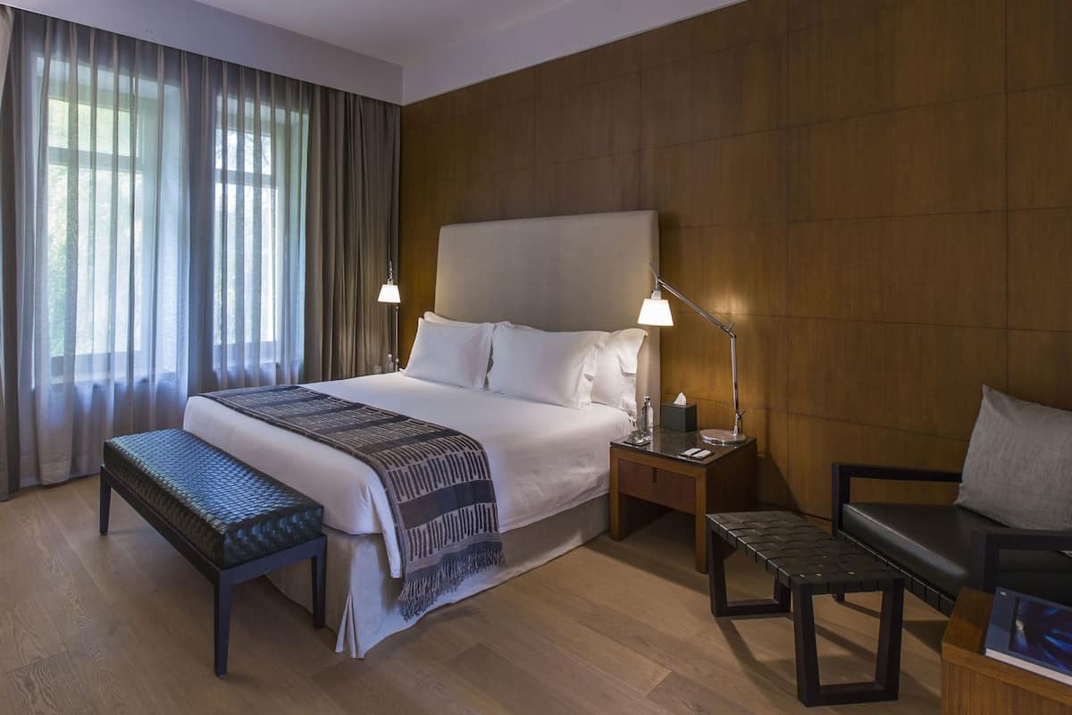 A hotel room with a large bed, bedside tables, a bench, a chair, and a wooden wall.