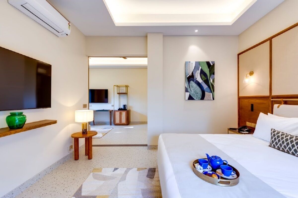 Une chambre d’hôtel avec un lit, une télévision, un tableau, un plateau avec des tasses et une théière, et une petite table avec une lampe.