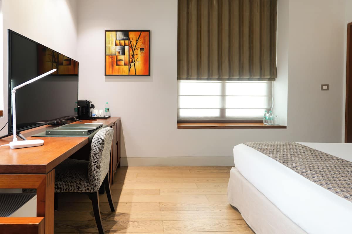 A hotel room with a wooden desk, a chair, a bed, a window with curtains, and a painting on the wall.