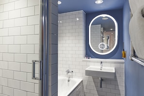 A modern bathroom with white tiled walls, a blue accent wall, a glass shower enclosure, a sink with a mirror, and a handheld soap dispenser.