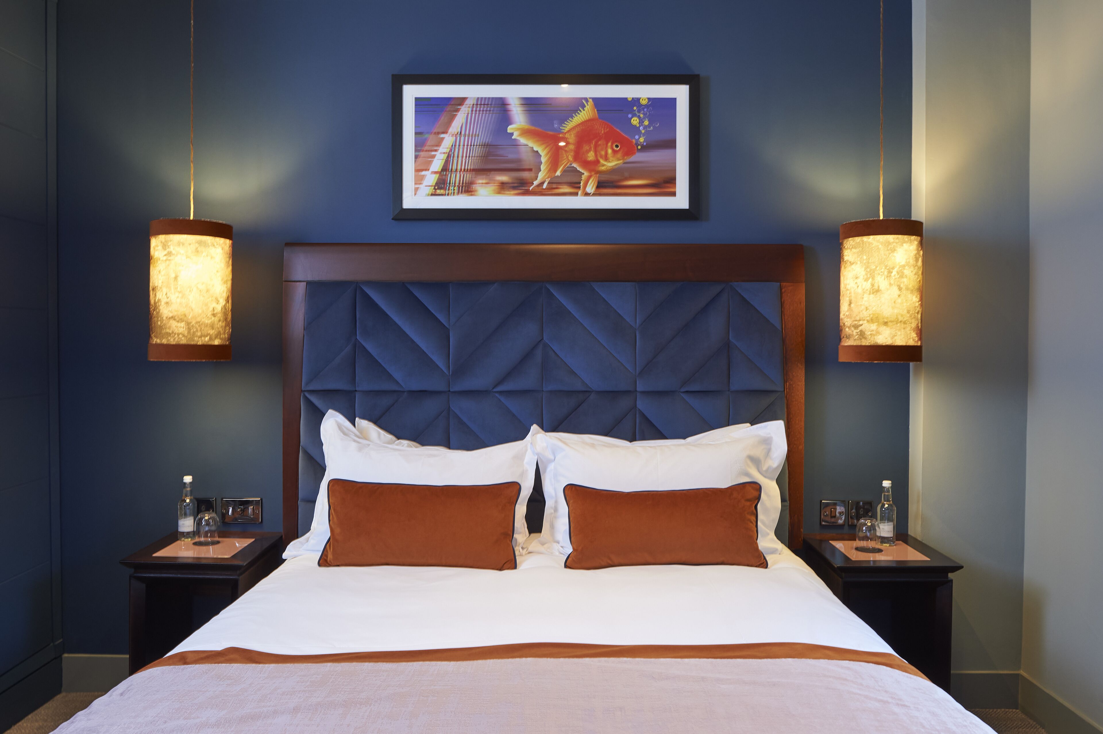 A hotel room with a large bed, two nightstands, a framed artwork above the bed, and two wall-mounted lamps.