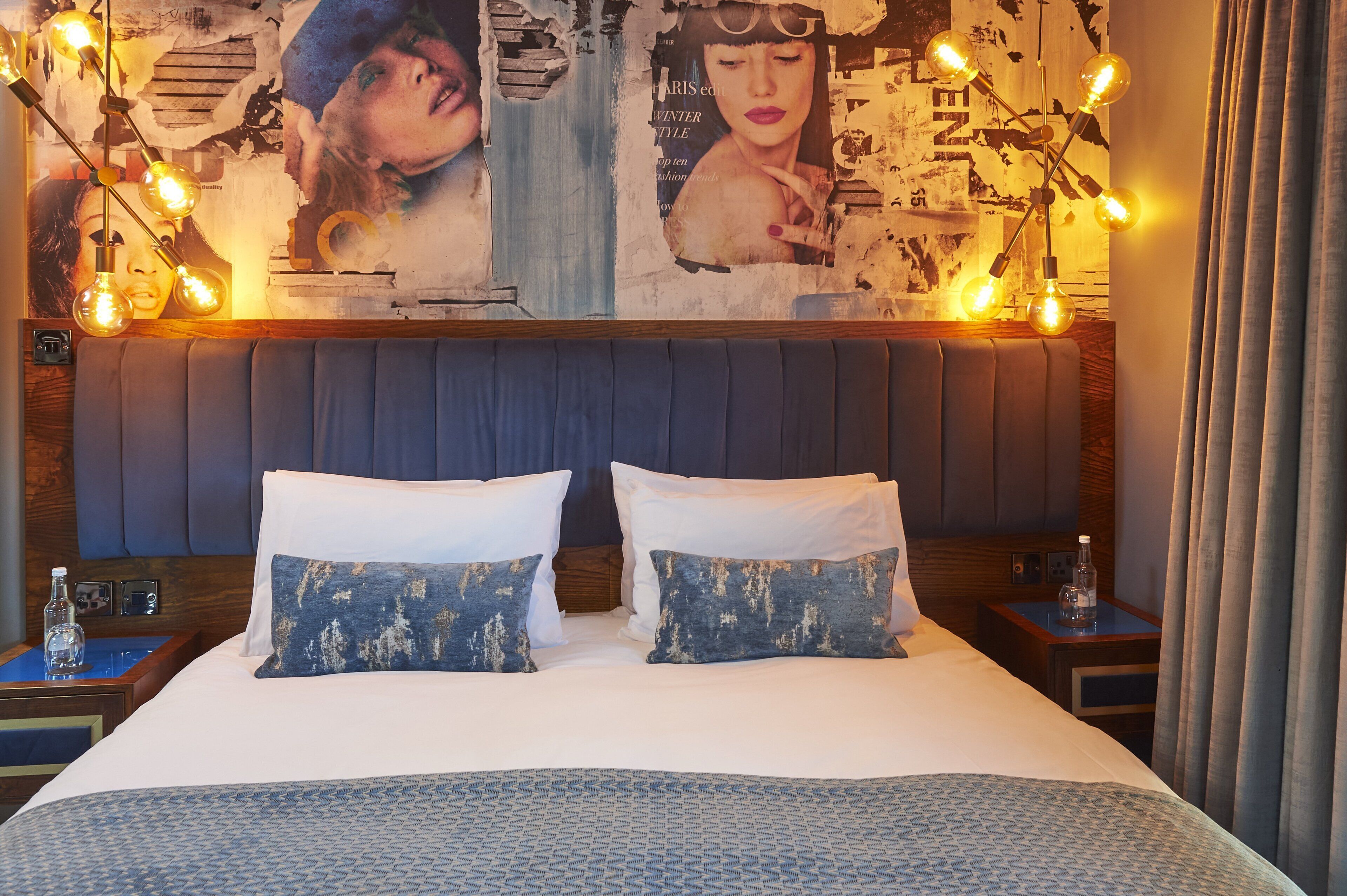 A neatly made bed with two pillows, a headboard, and a wall with posters and a light fixture.