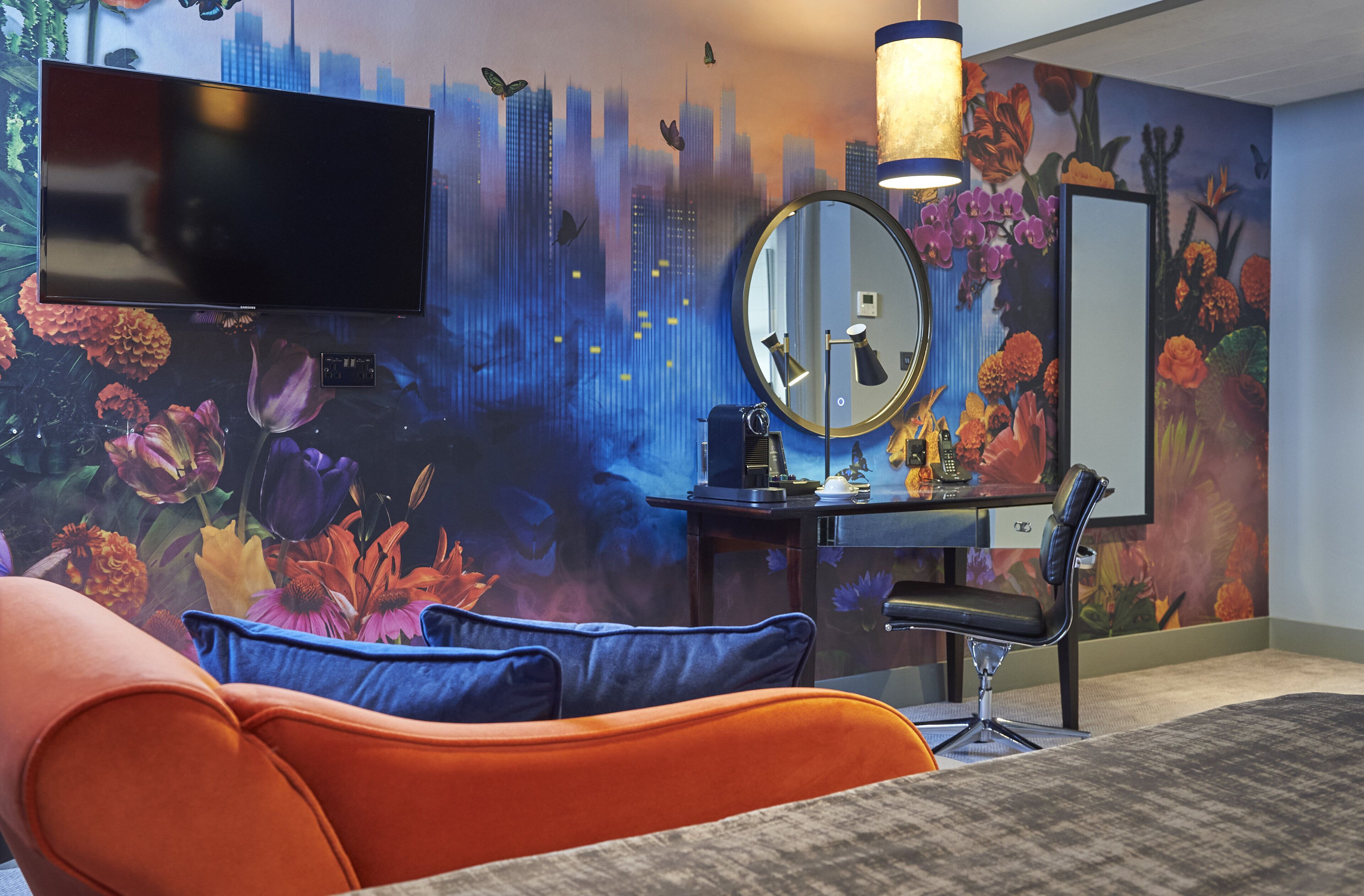 A modern bedroom with a cityscape mural, a flat-screen TV, a round mirror, a desk with a lamp, and an orange chair.
