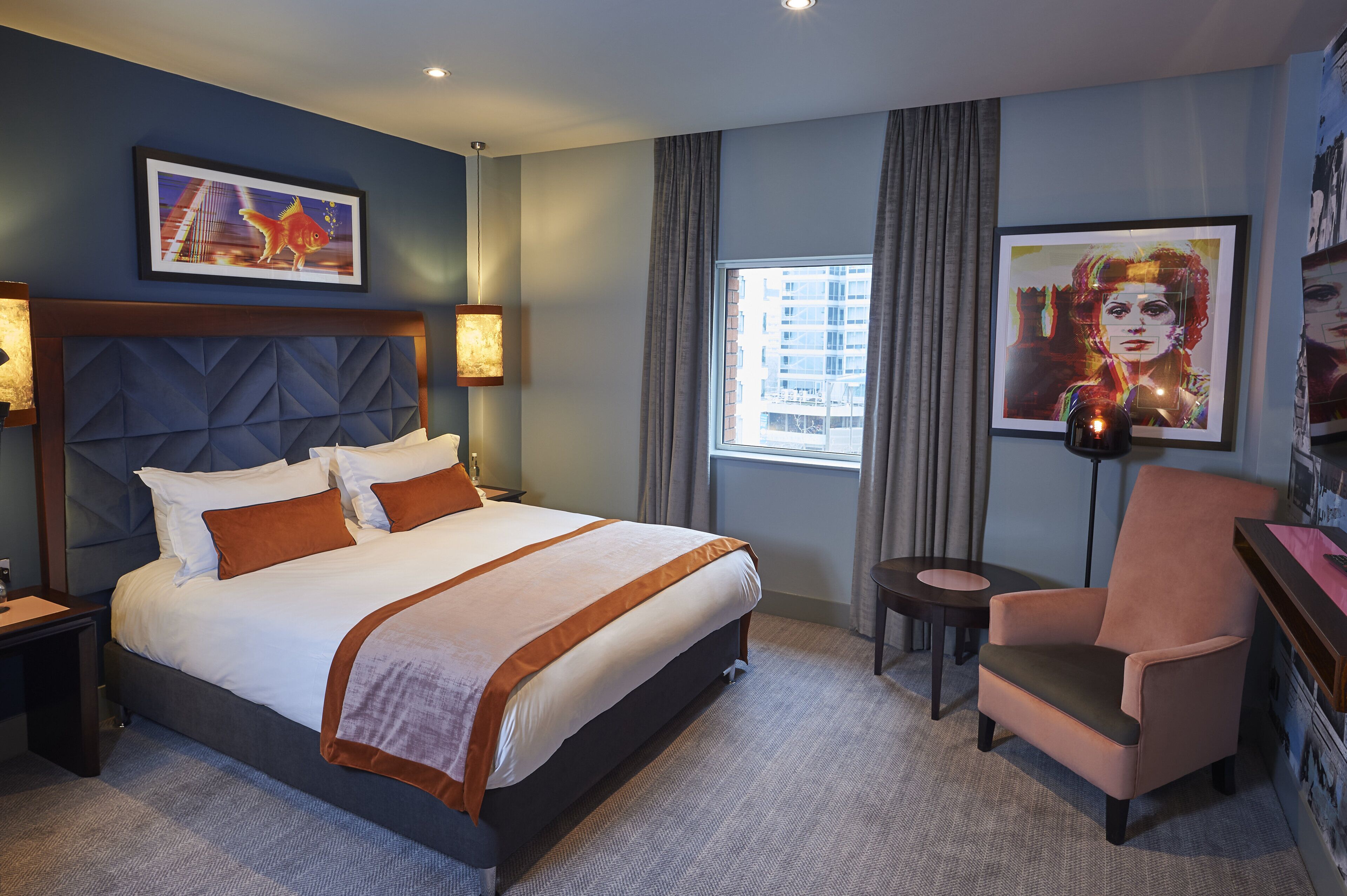 A hotel room with a large bed, a chair, a small table, and artwork on the walls.