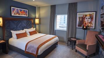 A hotel room with a large bed, a chair, a small table, and artwork on the walls.