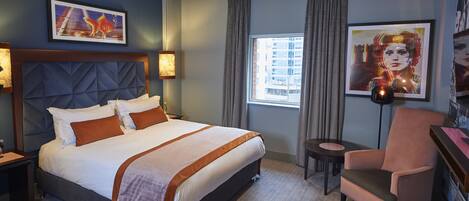 A hotel room with a large bed, a chair, a small table, and artwork on the walls.