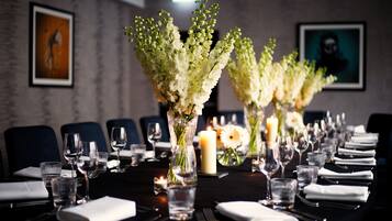 A long dining table set with glassware, plates, and napkins, flanked by vases with white flowers, in a room with framed artwork on the walls.