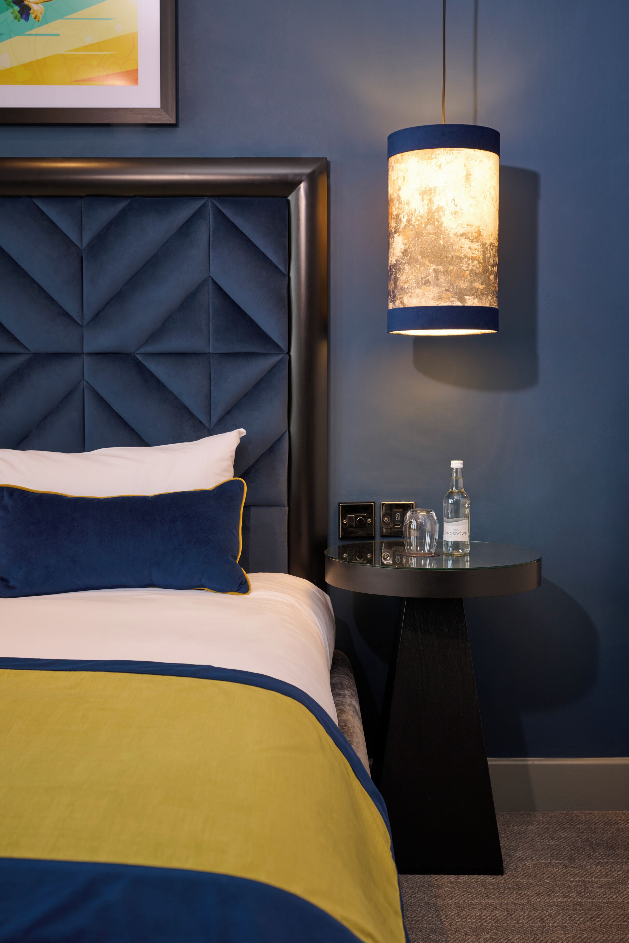 A neatly made bed with a blue and yellow bedspread, a white pillow, and a dark blue pillow. There is a bedside table with a bottle, glasses, and a small electronic device. The room has a blue wall and a textured headboard.