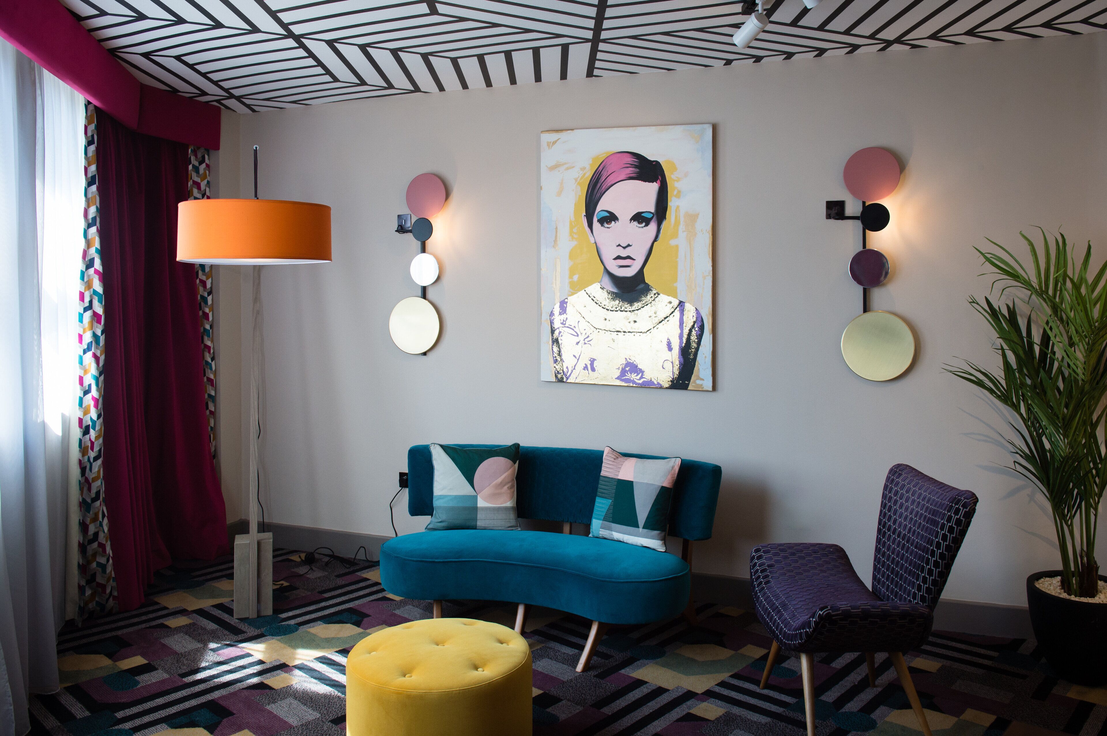 A modern living room with a teal sofa, a yellow ottoman, a purple chair, and a large painting on the wall.