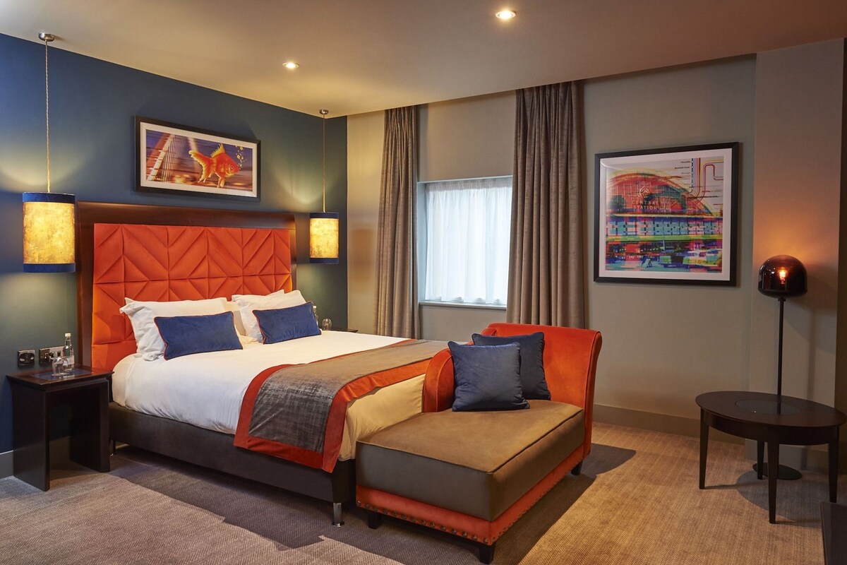 A hotel room with a large bed, a sofa, a desk, and a chair. There are two lamps, a window with curtains, and two framed pictures on the wall.