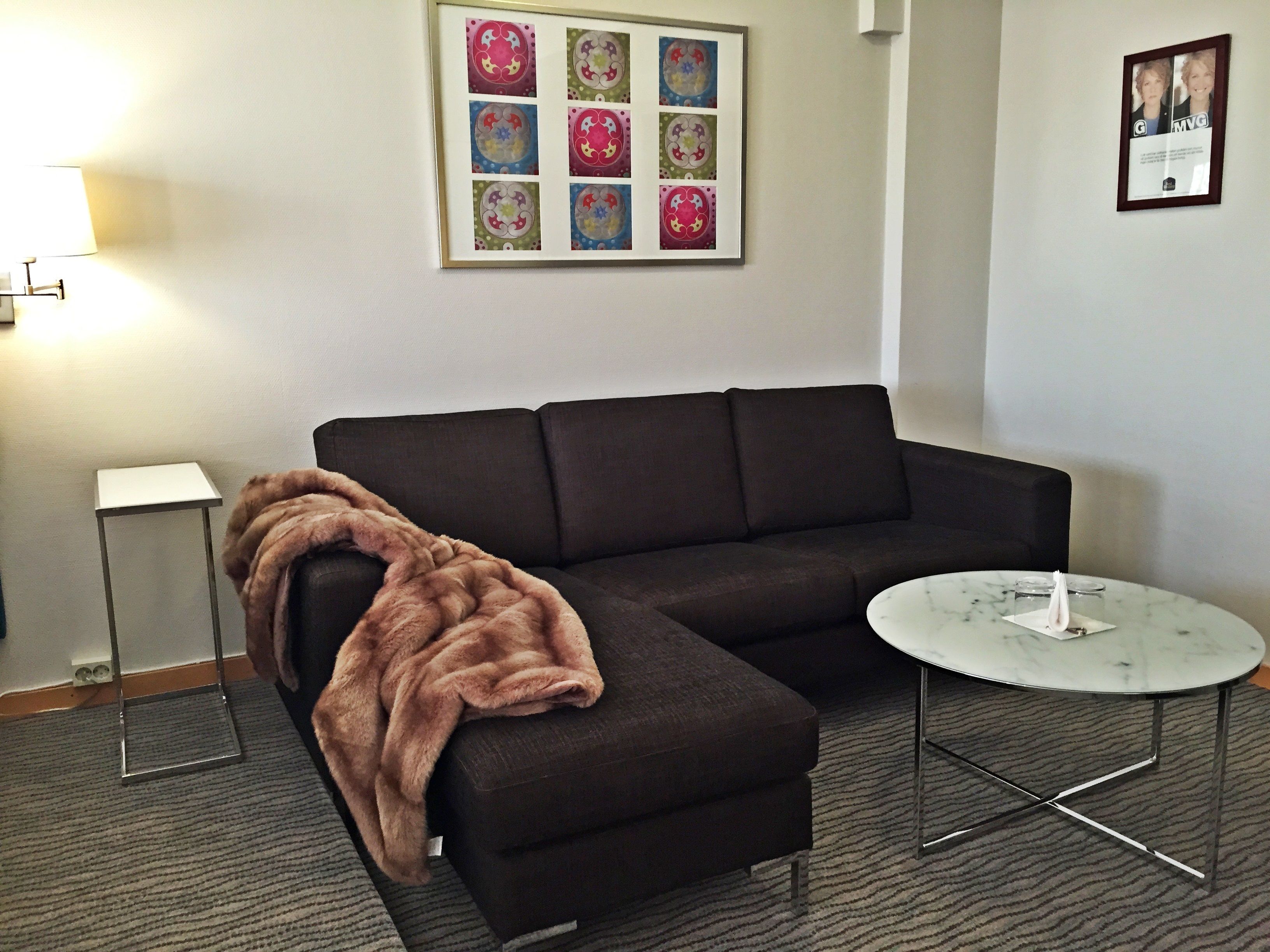 A living room with a dark brown sectional sofa, a small white side table, a round marble coffee table, and a framed picture on the wall.