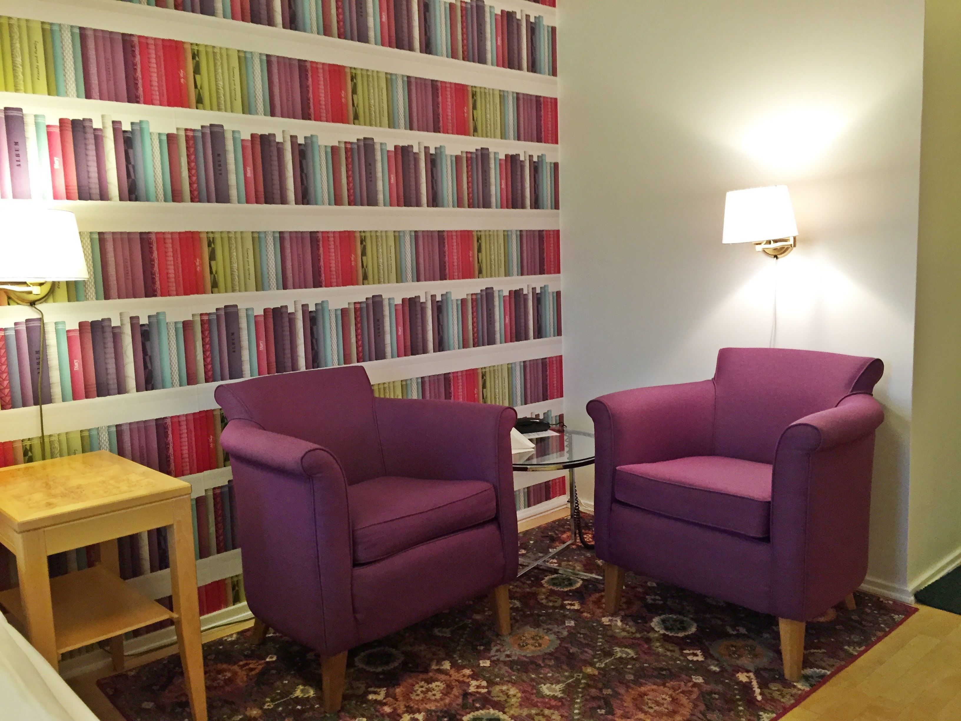 Una habitación con una estantería llena de libros, dos sillones morados, una mesita de madera y una alfombra estampada.