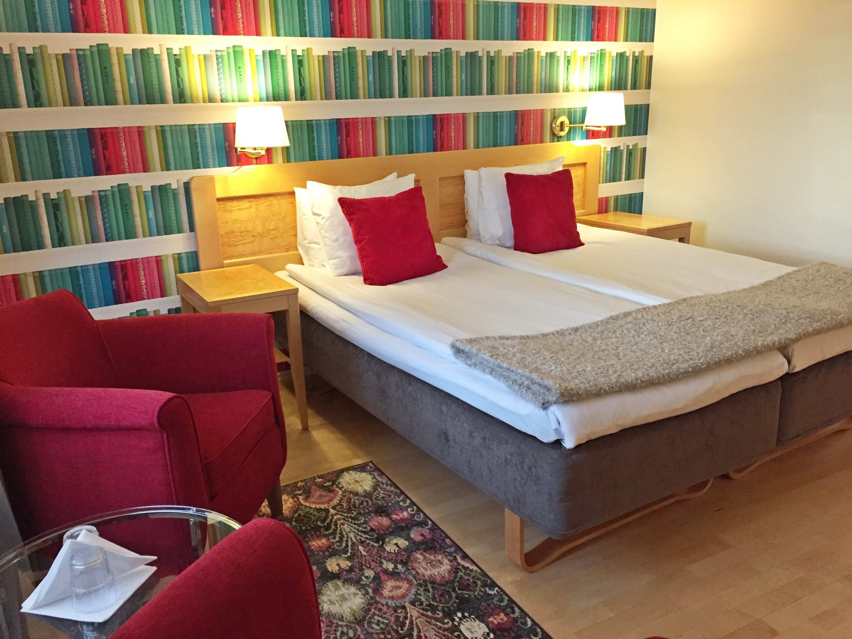 Una habitación de hotel con una cama grande, dos mesitas de noche, un sillón rojo, una pequeña mesa con un vaso, y una estantería llena de libros al fondo.