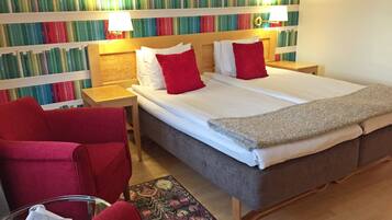 Una habitación de hotel con una cama grande, dos mesitas de noche, un sillón rojo, una pequeña mesa con un vaso, y una estantería llena de libros al fondo.