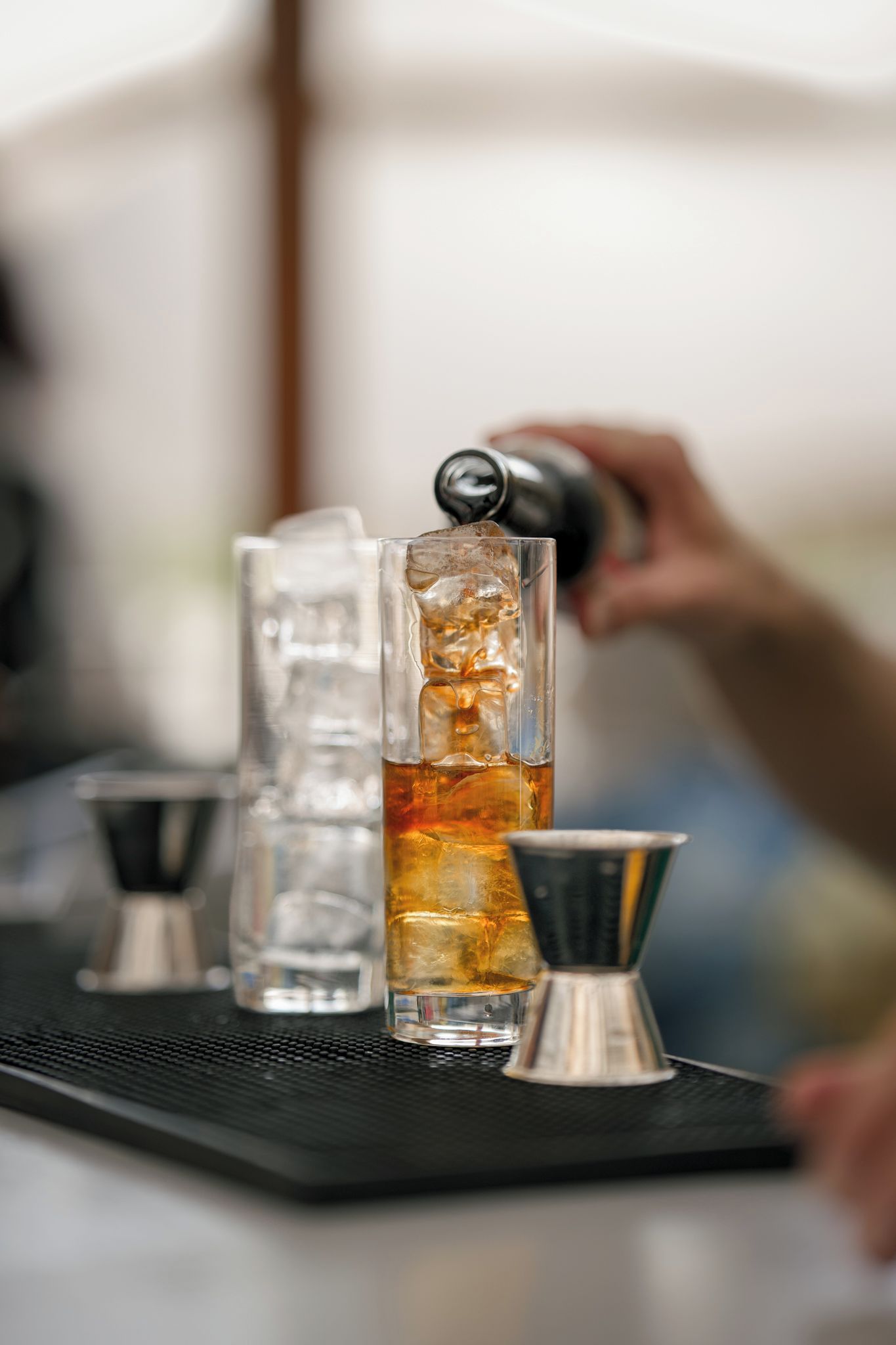 A bar setting with a tall glass filled with a golden liquid, ice cubes, and a metal jigger.