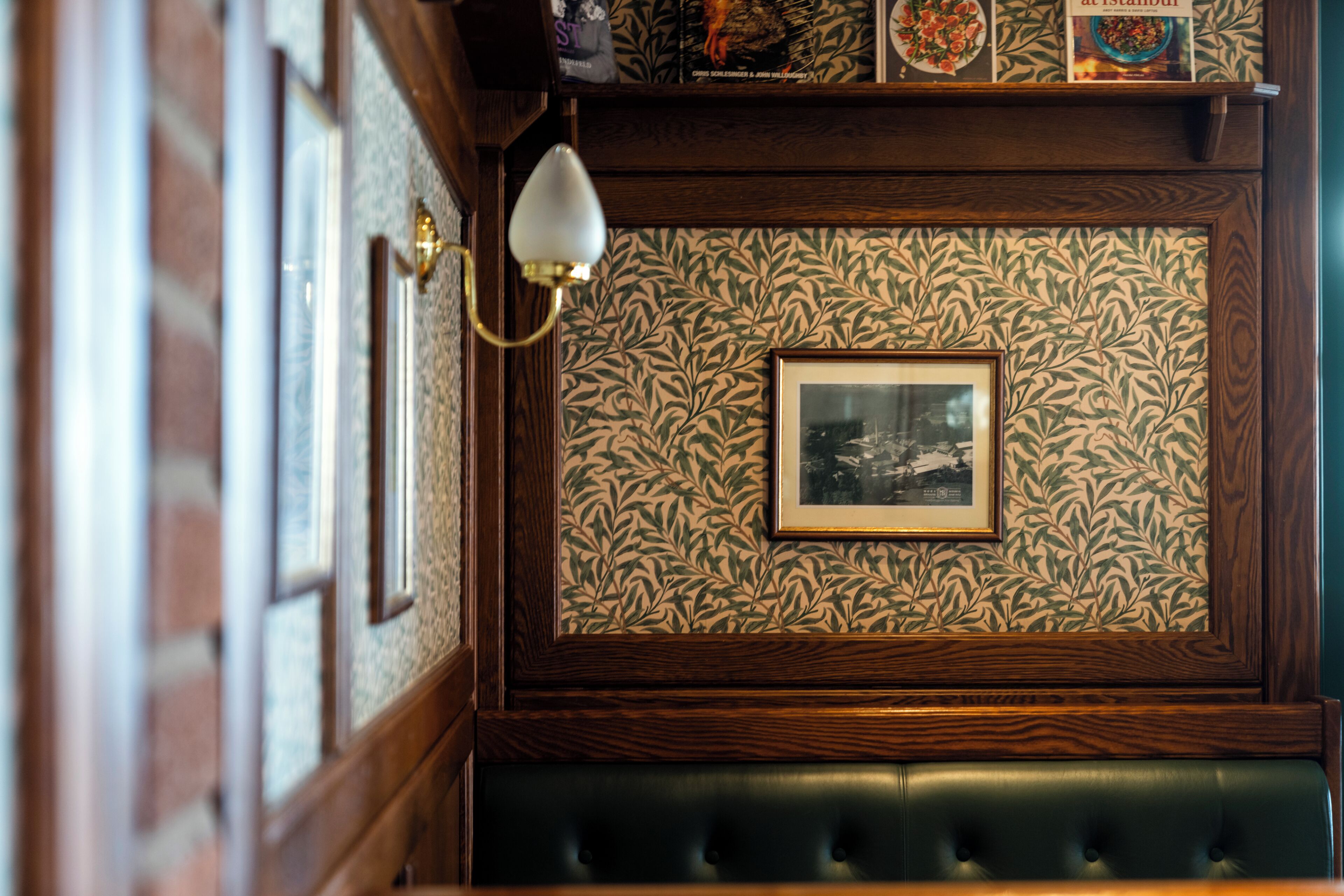 A room with a patterned wallpaper, a framed picture, a green upholstered bench, and a wall-mounted light fixture.
