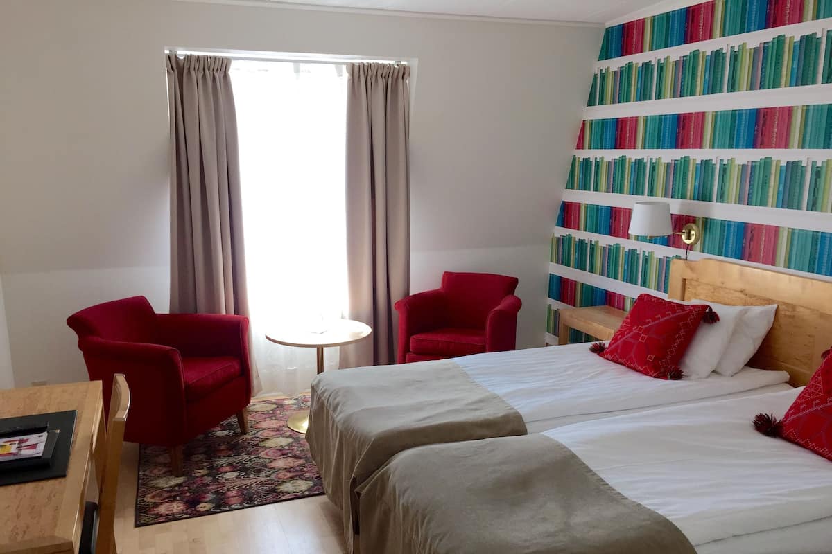 Une chambre d’hôtel avec un lit, deux fauteuils rouges, une petite table ronde et une bibliothèque remplie de livres aux couleurs vives.