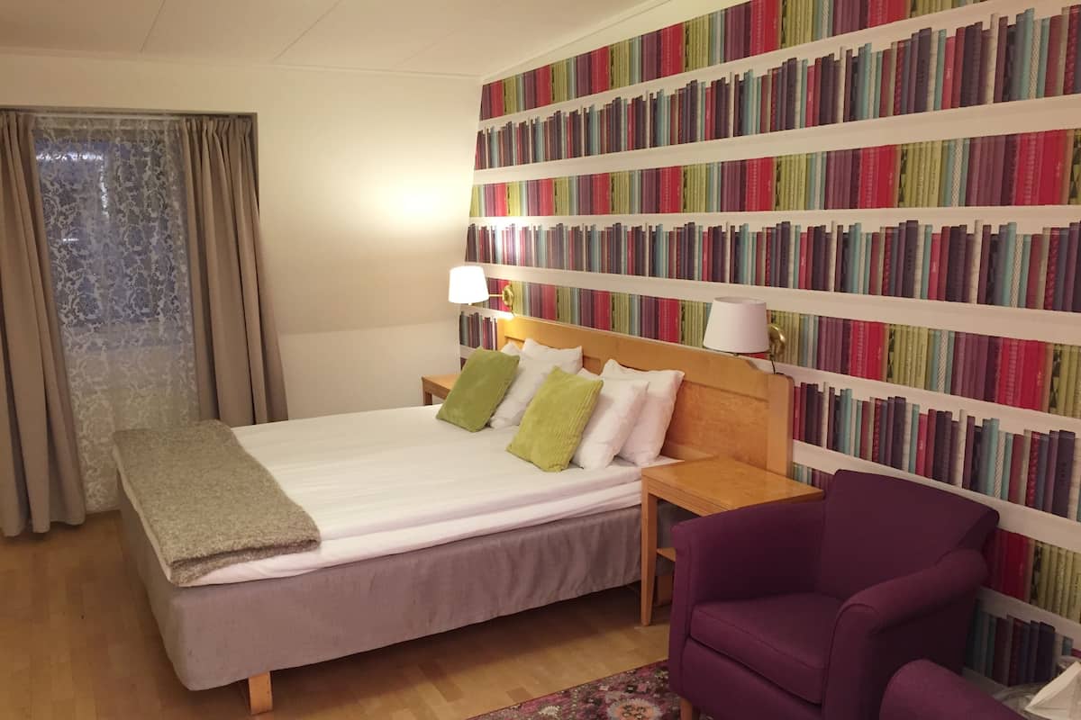 Une chambre d’hôtel avec un lit, un fauteuil violet, une bibliothèque et une fenêtre avec des rideaux.