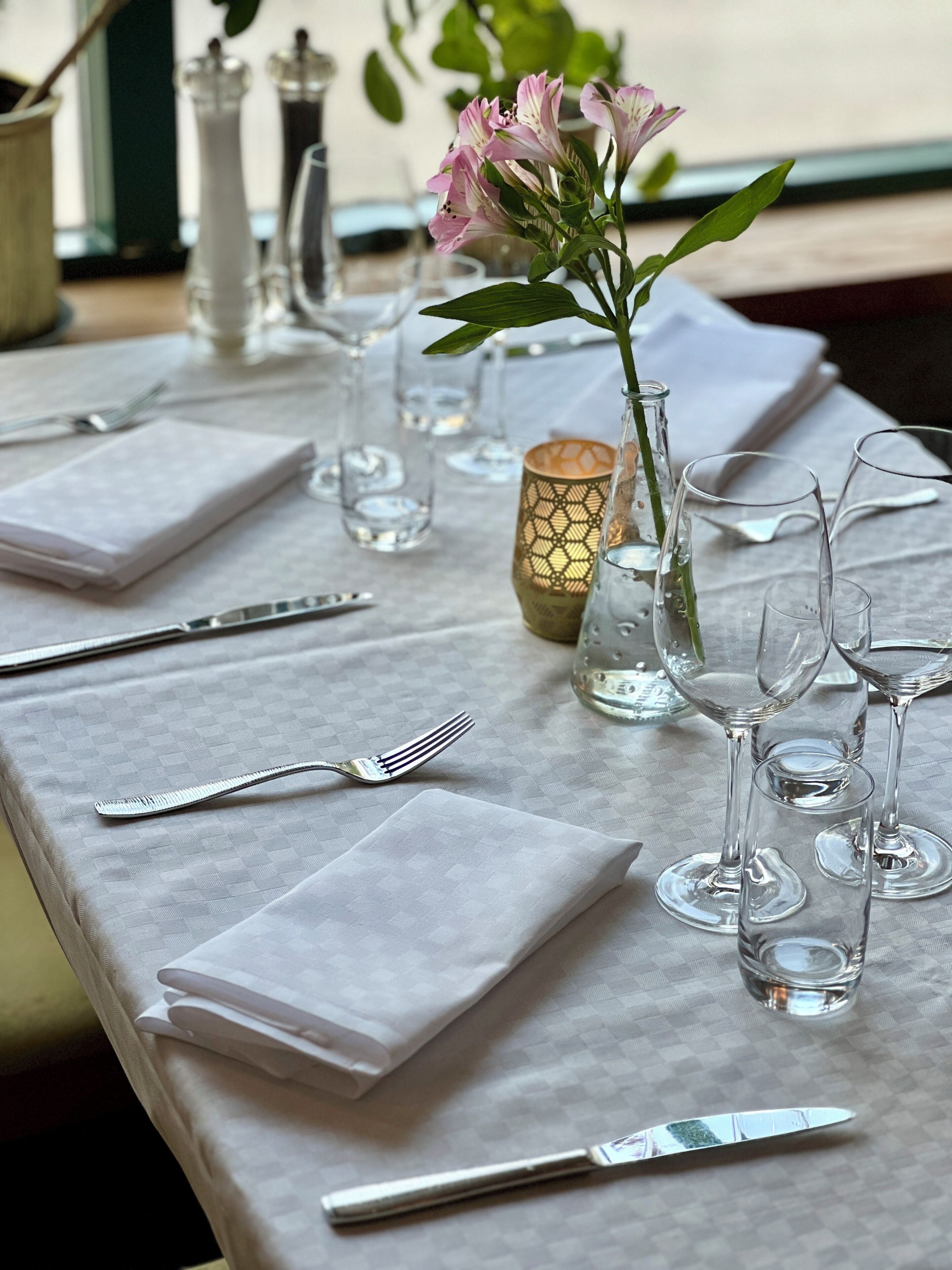 Uma mesa posta com copos de vidro transparente, um vaso com flores e uma toalha de mesa estampada.