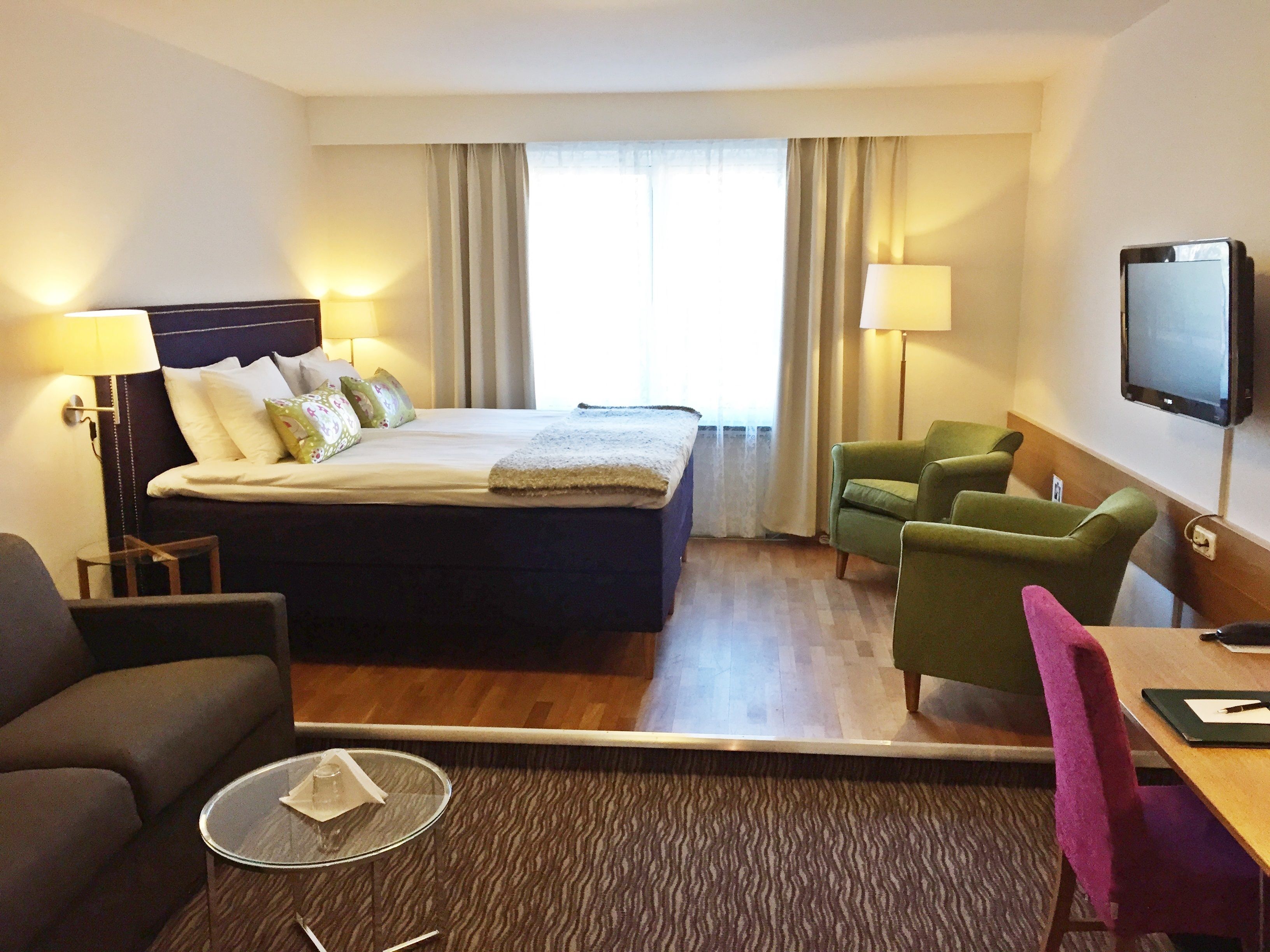 A hotel room with a bed, two chairs, a small table, a TV, and a window with curtains.