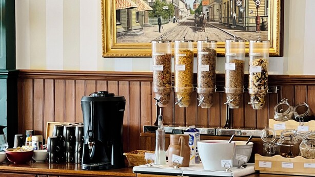 A breakfast buffet with various food items, coffee machines, and a painting of a street scene on the wall.