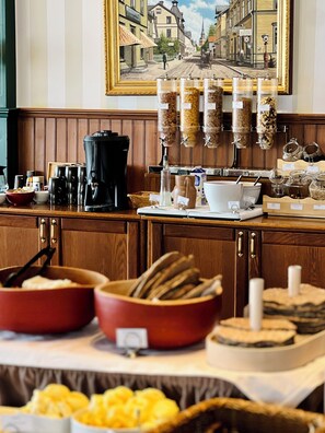 A breakfast buffet with various food items, coffee machines, and a painting of a street scene on the wall.