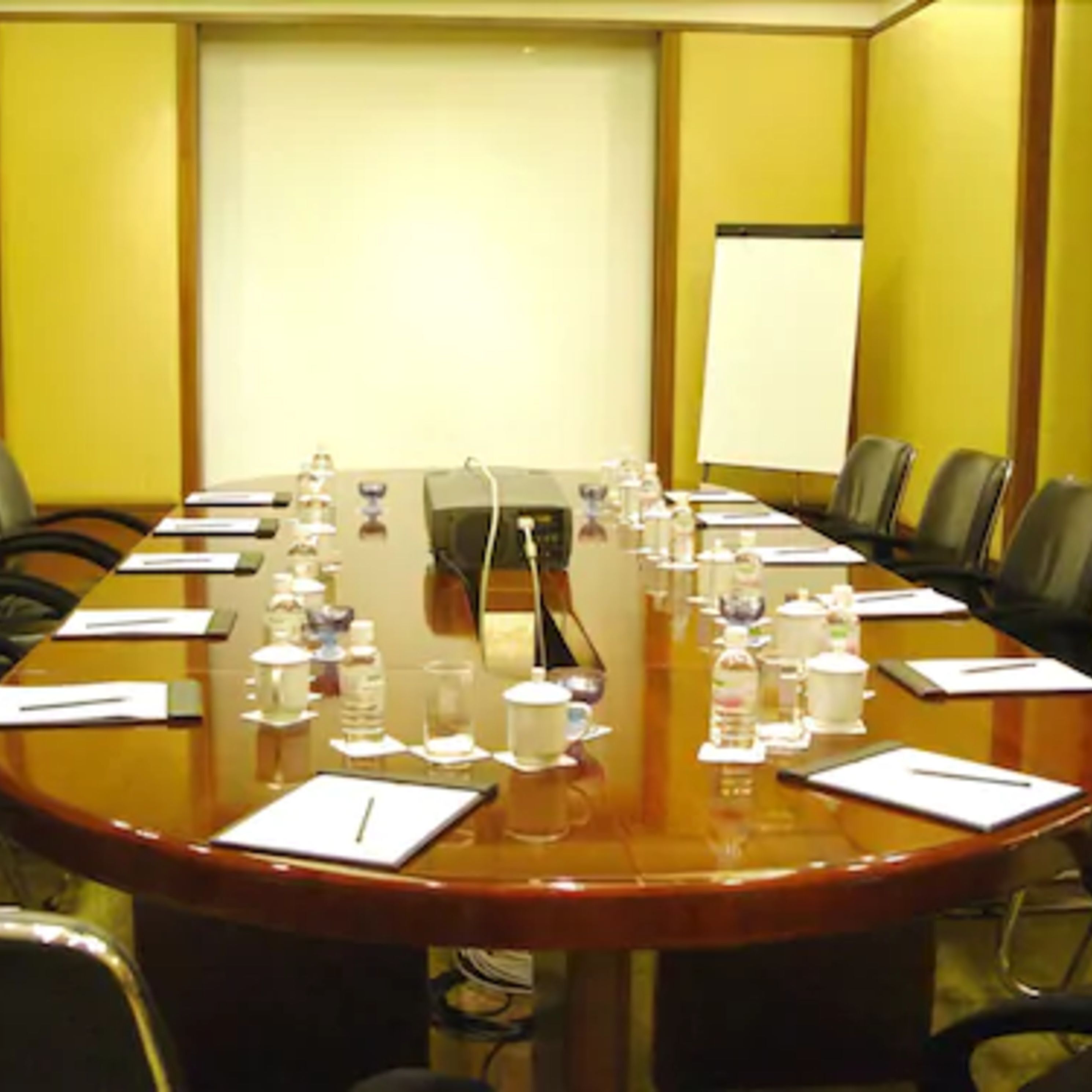 Un salón de conferencias con una larga mesa, sillas, tarjetas de identificación, botellas de agua y una pantalla de proyector.