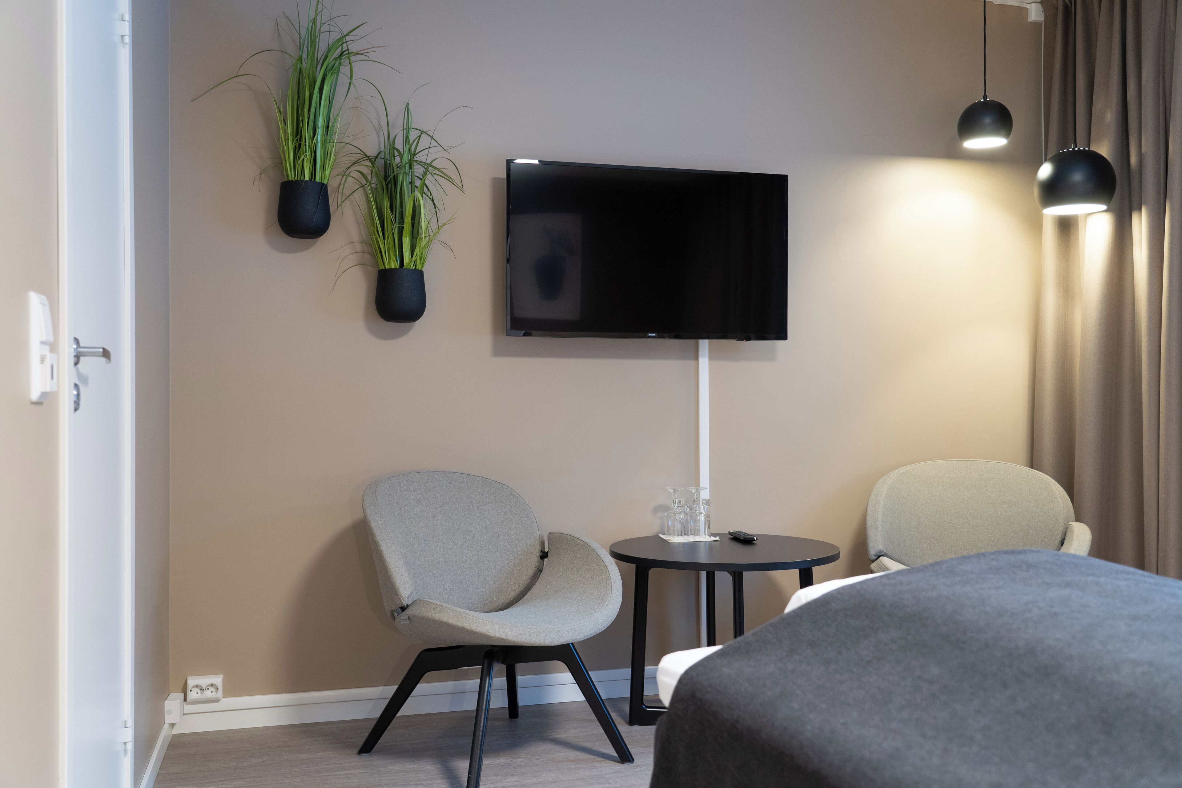 Una habitación de hotel moderna con un televisor de pantalla plana en la pared, dos sillones, una mesita con un vaso y dos lámparas colgantes.