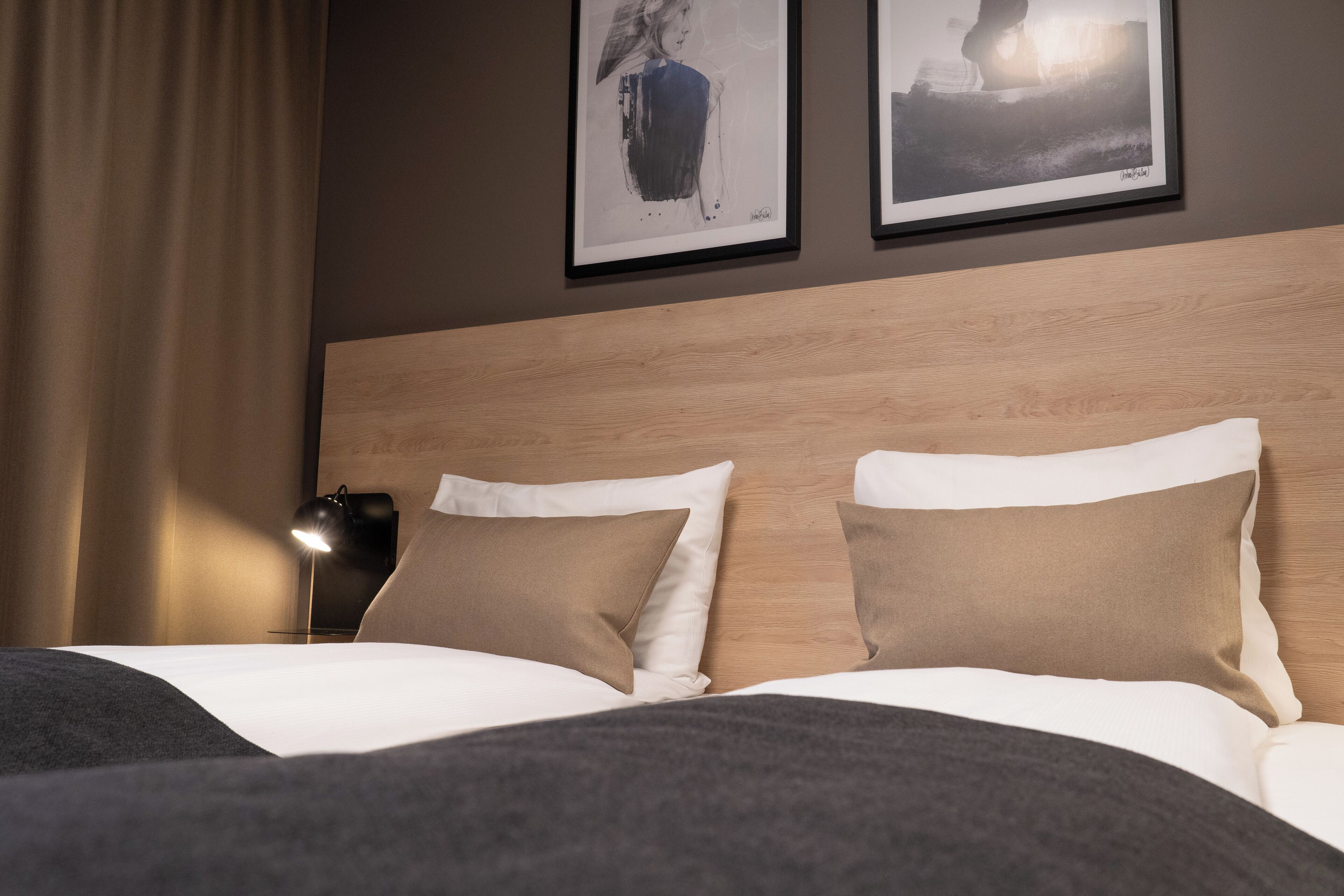 Habitación de hotel con cabecera de madera, dos almohadas, una lámpara negra y dos cuadros en la pared.
