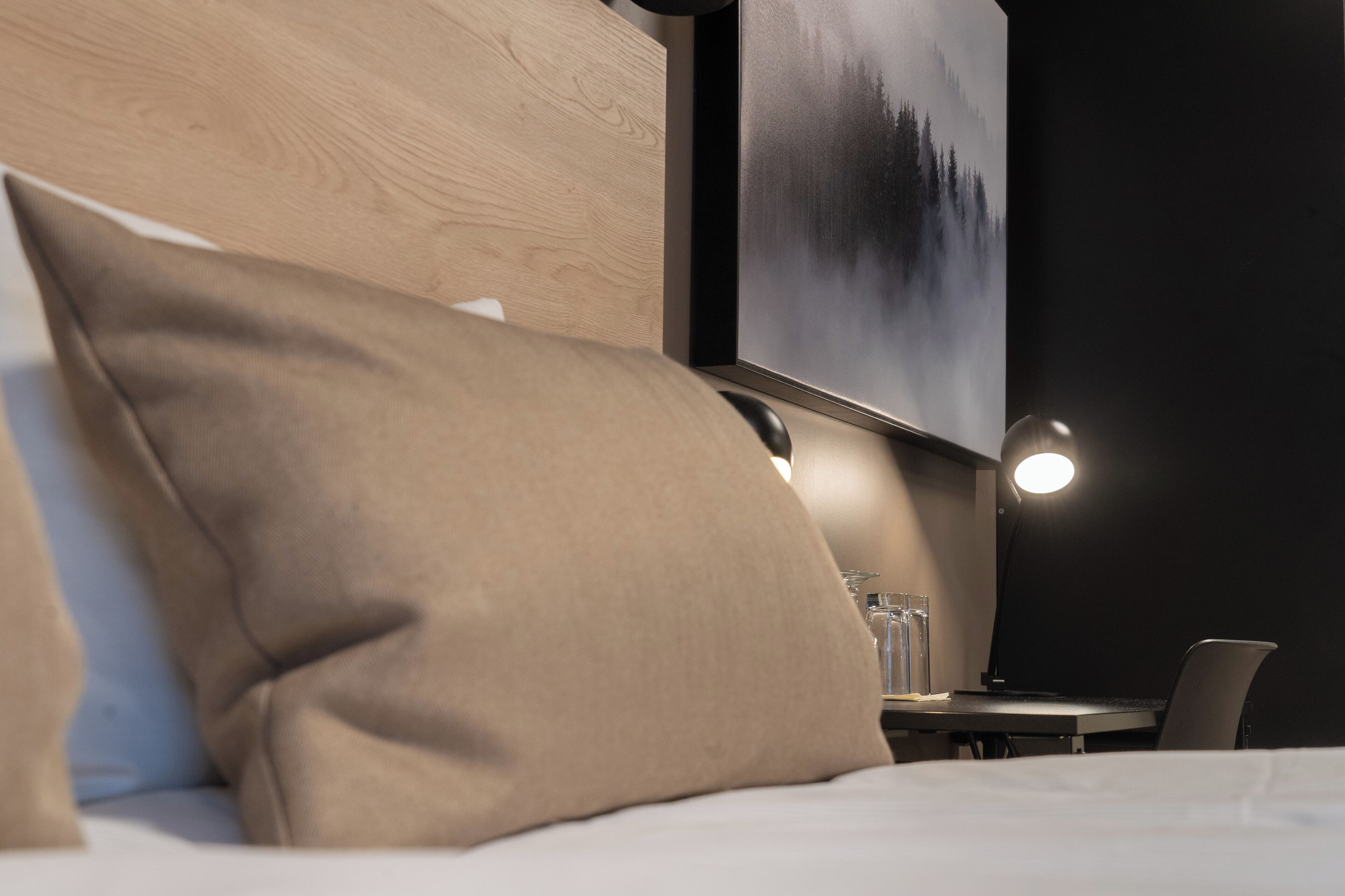 A hotel room with a bed, a desk, and a chair. There is a painting on the wall and a lamp on the desk.